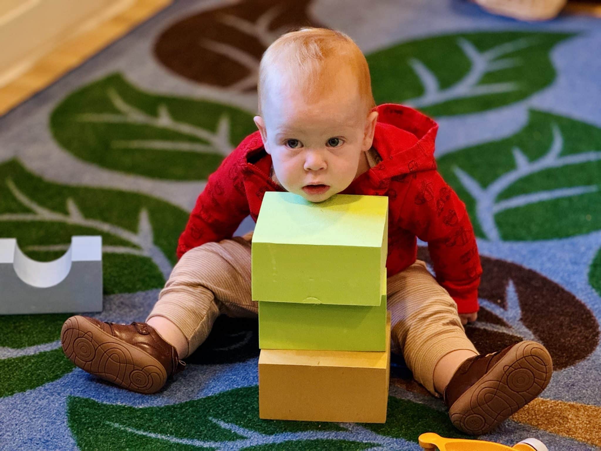 The image shows a young child engaged in play on a colorful, leaf-patterned rug. The child, who has a focused